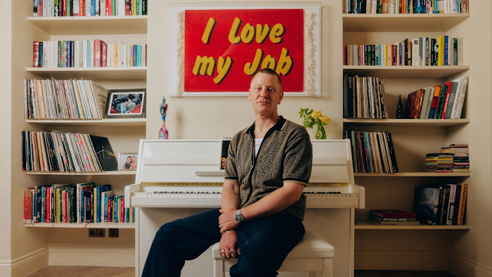 책이 늘어선 방에서 ‘I Love My Job’이라고 적힌 액자 아래 흰색 피아노 앞에 앉아 있는 남성의 초상.