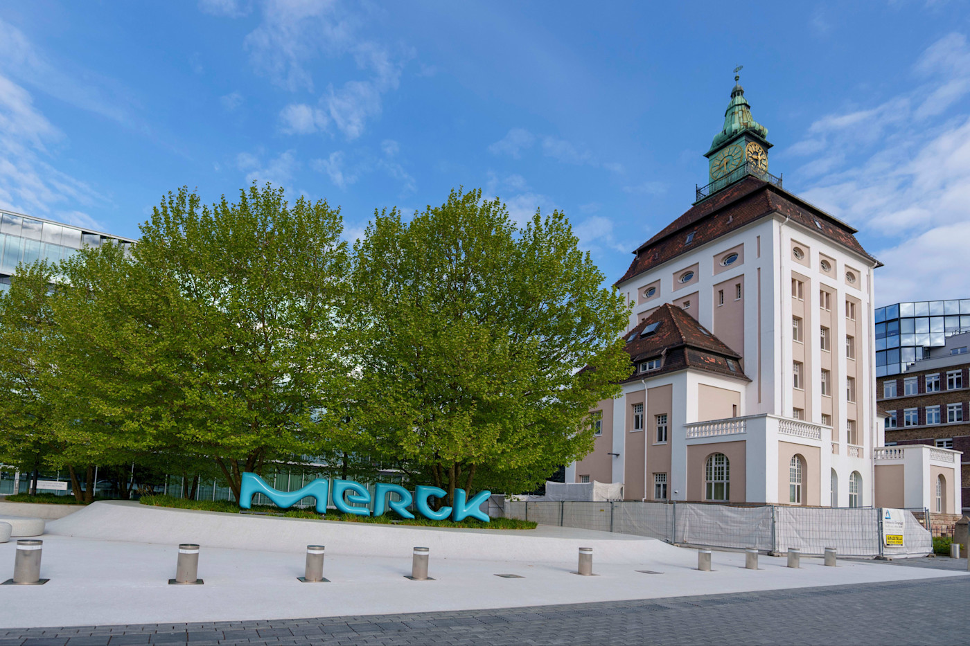 역사적인 P'tzer 타워가 Merck 본사 옆에 서 있으며, 앞쪽에 나무와 커다란 파란색 Merck 간판이 보인다.