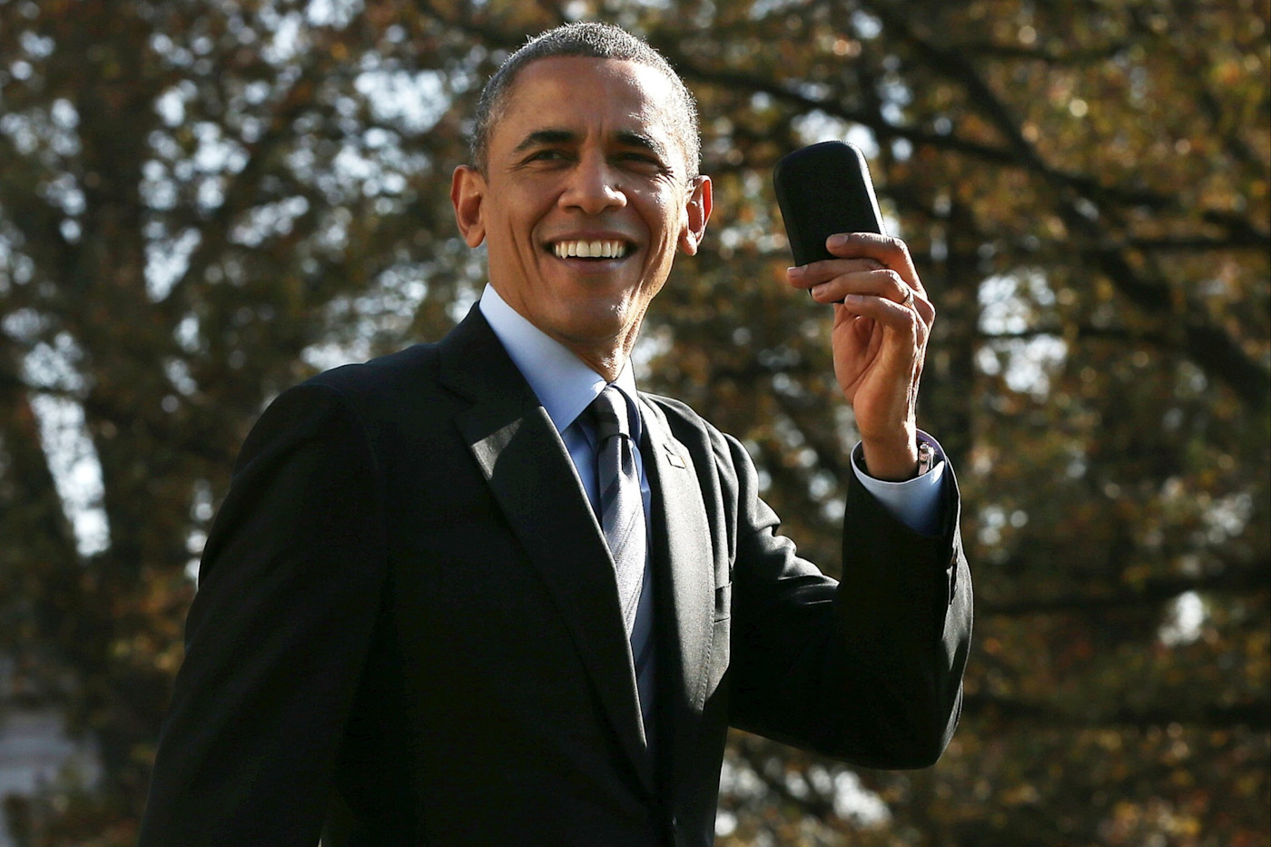 Barack Obama가 정장과 넥타이를 착용한 채 야외에서 미소 지으며 BlackBerry 휴대전화를 들어 보이고 있다.