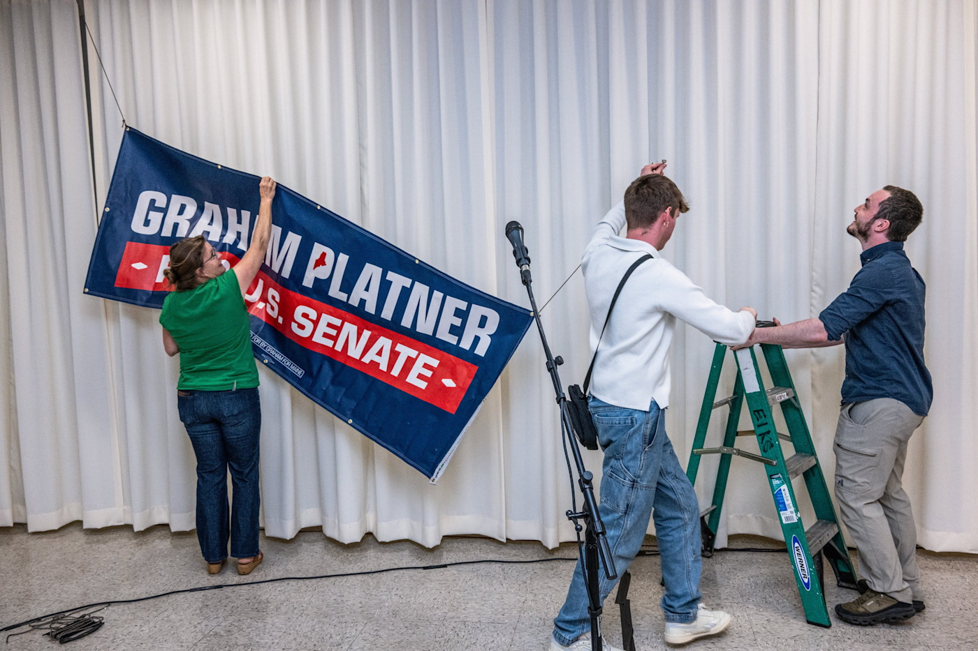 세 명의 선거운동원이 타운홀 행사를 준비하며 흰 커튼 앞에 ‘Graham Platner U.S. Senate’ 표지판을 걸고 있다.