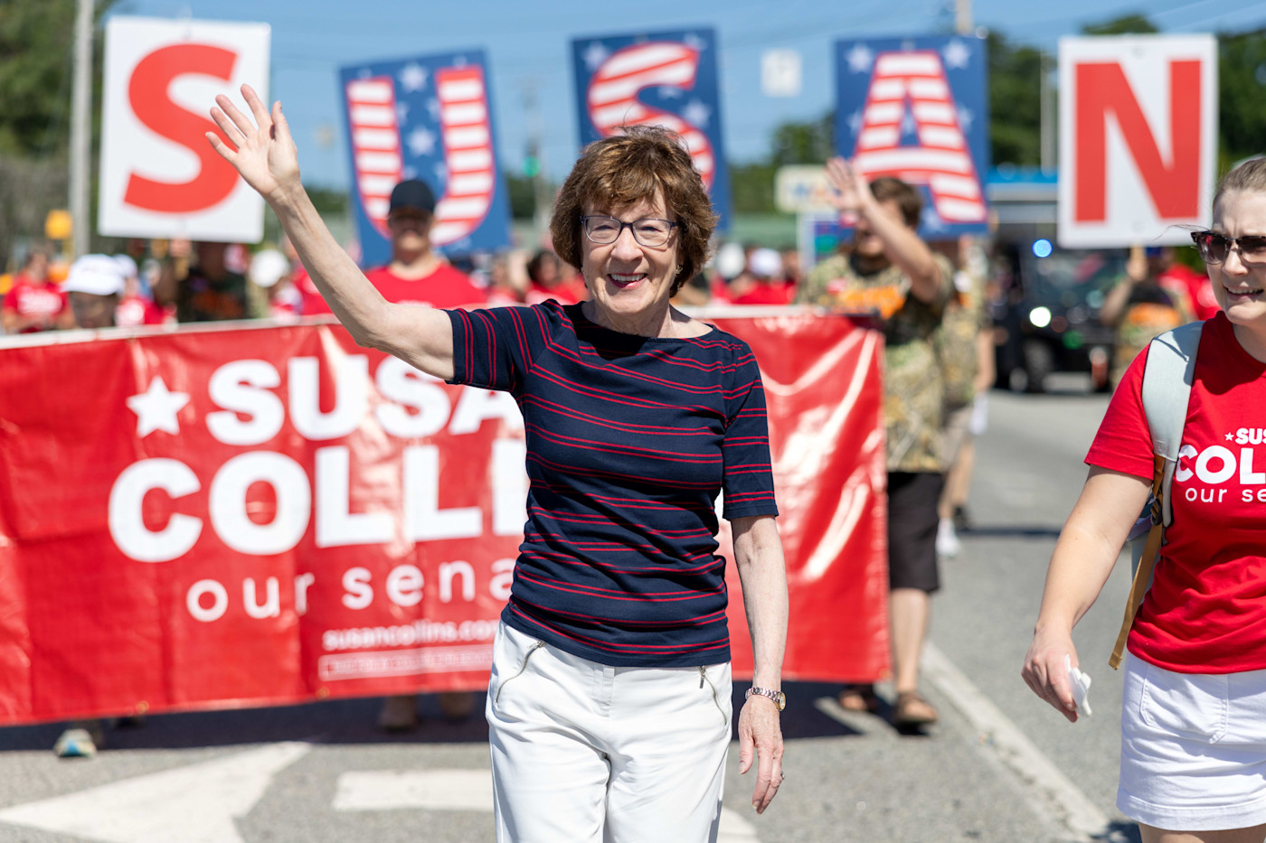 Susan Collins가 Moxie Day Parade에서 손을 흔들며 걷고 있고, 뒤에는 지지자들이 그녀의 이름이 적힌 팻말과 현수막을 들고 있다.