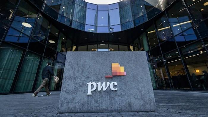 현대적인 유리 오피스 빌딩 앞 대형 석재 표지판에 있는 PwC 로고와 지나가는 사람.