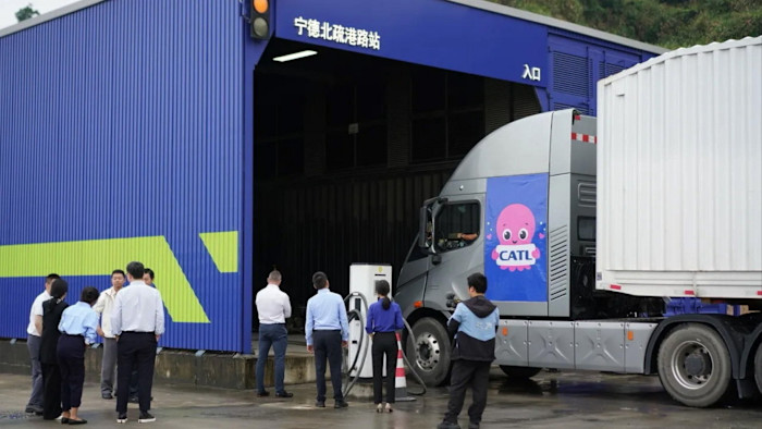 CATL 로고가 부착된 전기 화물트럭이 배터리 교환 허브로 진입하고, 여러 사람이 시설 밖에서 지켜보고 있다.
