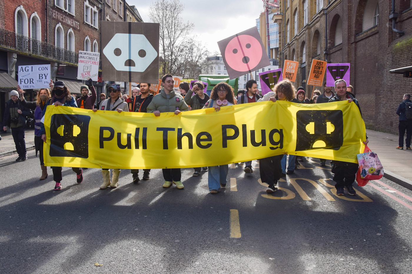 시위대가 플러그 얼굴 아이콘이 그려진 커다란 노란색 ‘Pull The Plug’ 현수막을 들고, 규제되지 않은 AI와 데이터센터에 반대하는 팻말을 들고 행진하고 있다.