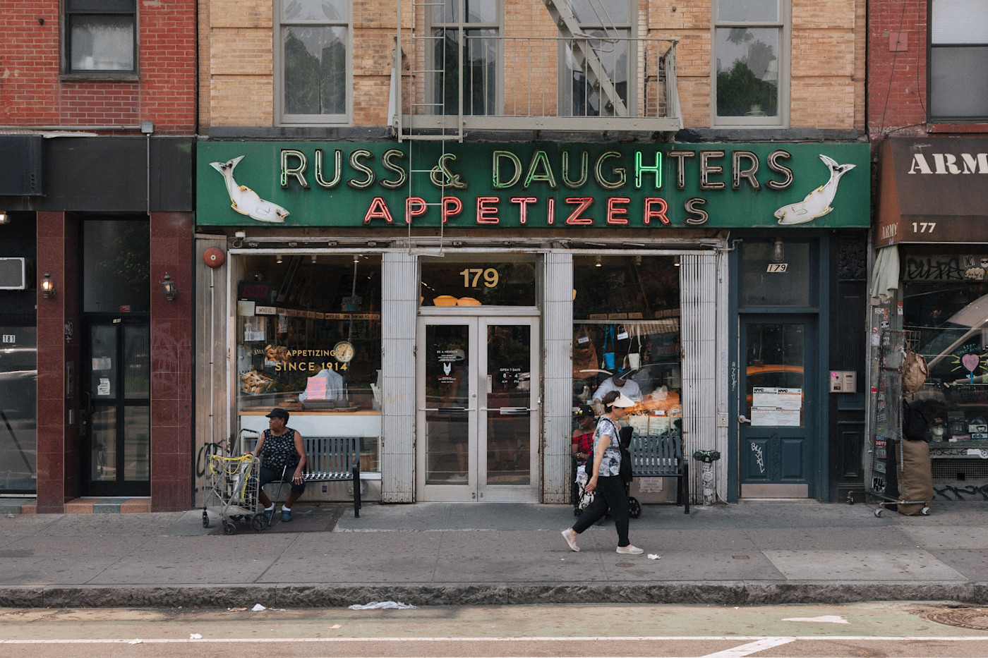 네온 간판이 있는 Russ & Daughters Appetizers 매장 앞, 보행기를 곁에 둔 채 벤치에 앉아 있는 여성과 지나가는 또 다른 여성.