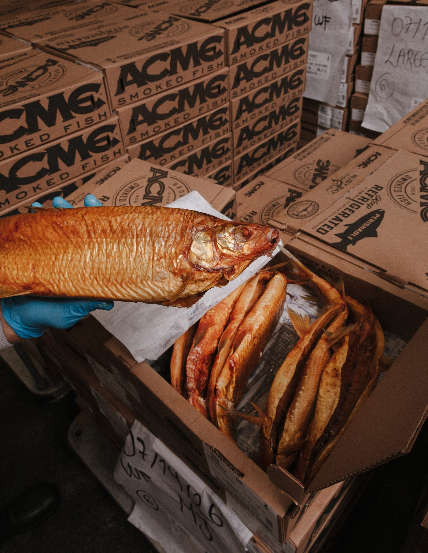 파란 장갑을 낀 사람이 훈제 생선이 담긴 상자 위로 훈제 생선을 들고 있으며, 주변에는 ACME Smoked Fish 상자가 쌓여 있다.