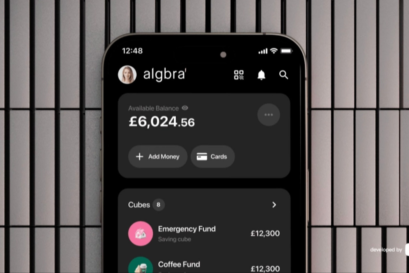Algbra 뱅킹 앱이 표시된 스마트폰 화면에 이용 가능 잔액 £6,024.56와 ‘Emergency Fund’, ‘Coffee Fund’로 표시된 저축 항목이 보인다.