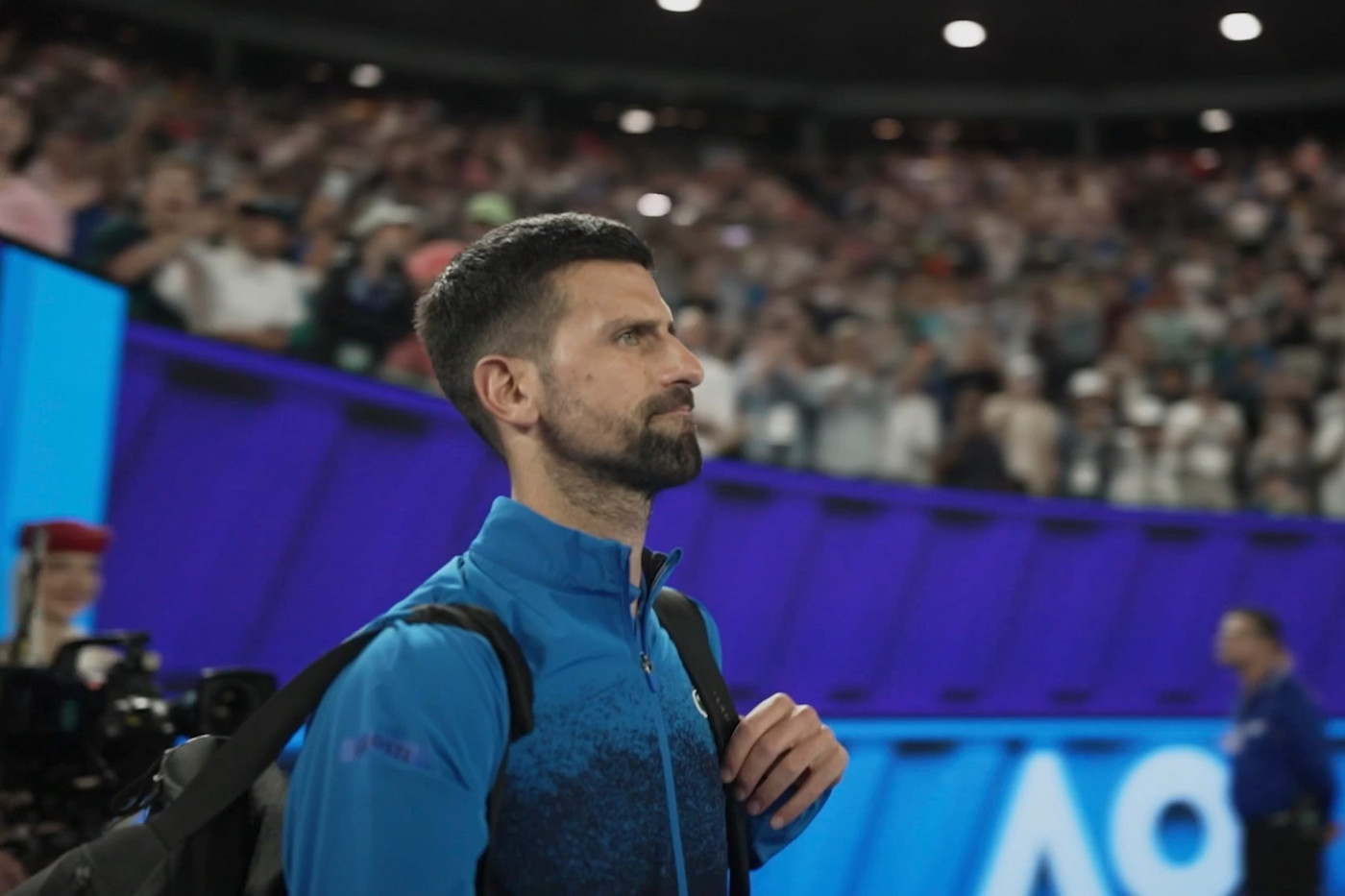 Novak Djokovic가 파란 재킷을 입고 코트 옆에 서서 집중한 표정을 짓고 있으며, 뒤로 관중으로 가득 찬 경기장이 보인다.