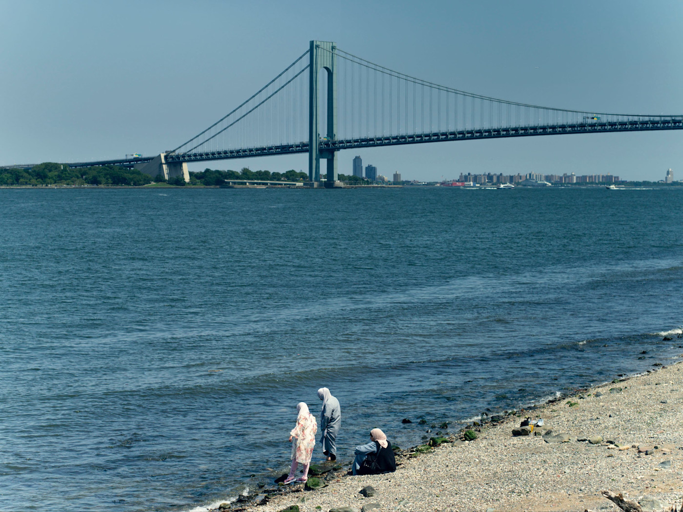 Buono Beach의 바위 해안에 세 명이 서 있고 한 명이 앉아 있으며, 뒤로 물 위를 가로지르는 Verrazzano-Narrows Bridge가 보인다.