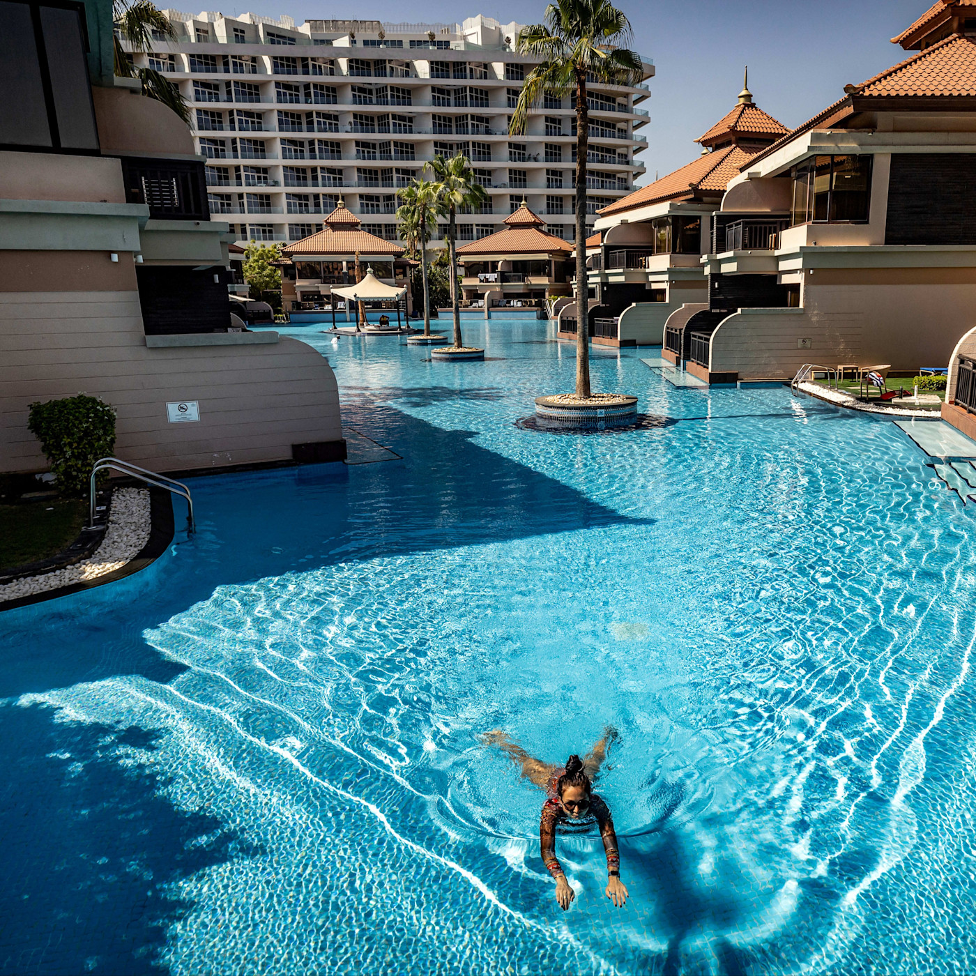 여성이 Anantara The Palm Dubai Resort의 야자수와 리조트 건물로 둘러싸인 햇살 가득한 대형 수영장에서 수영하고 있다.