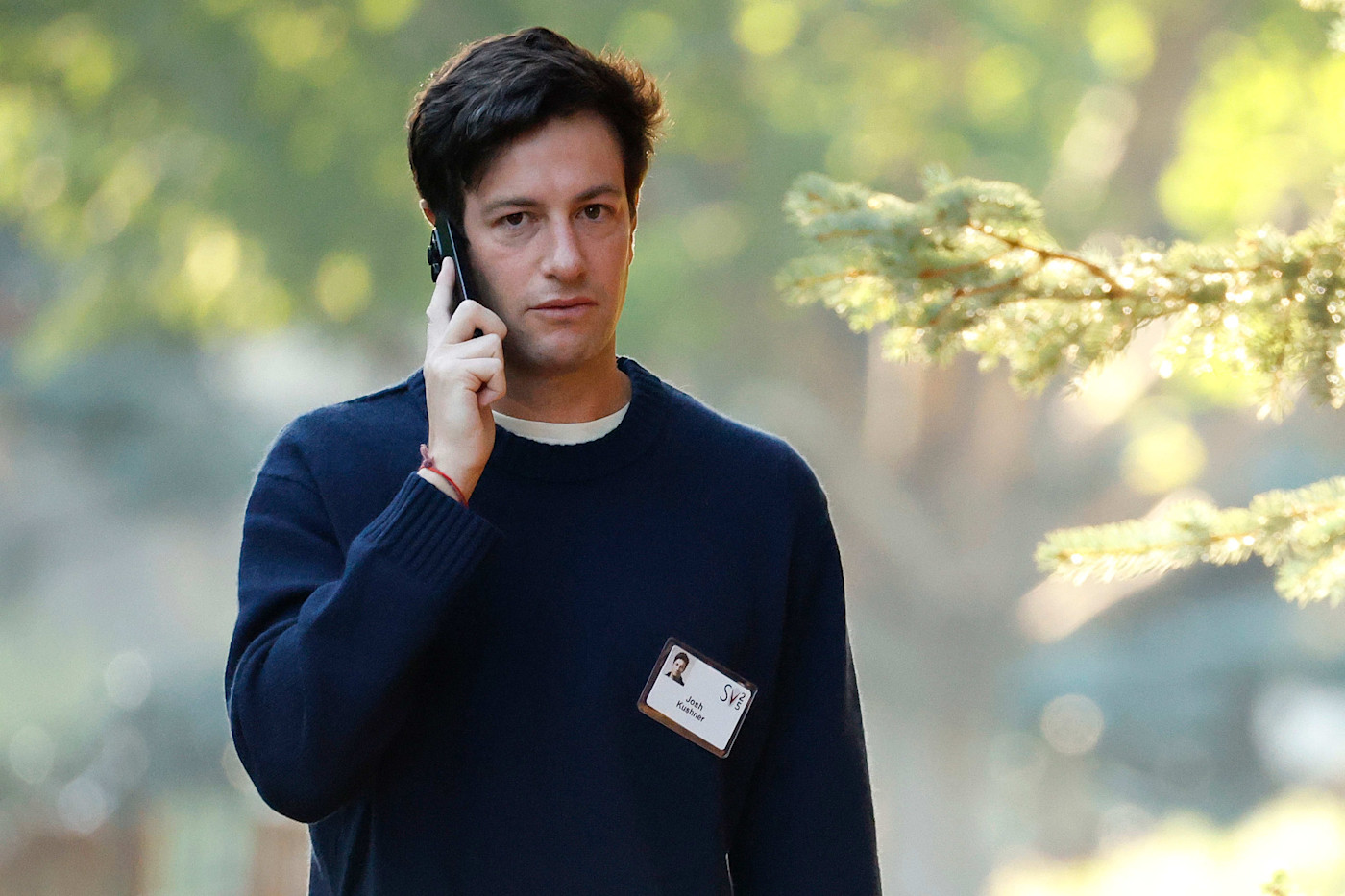 Josh Kushner가 어두운색 스웨터와 컨퍼런스 배지를 착용하고 야외를 걸으며 휴대전화로 통화하고 있다.