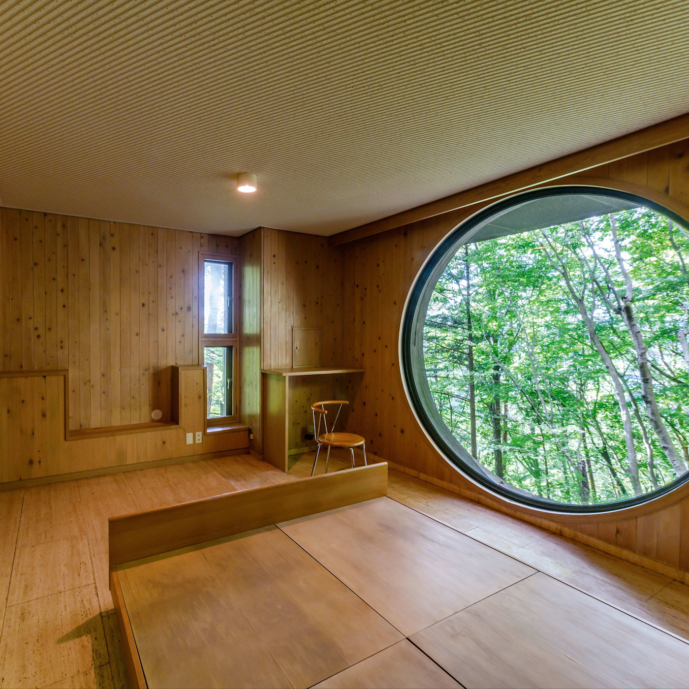 나무 패널로 꾸민 Capsule House K 내부. 큰 원형 창문 너머로 나무가 보이고, 낮은 플랫폼 침대와 의자가 놓인 작은 책상이 있다.