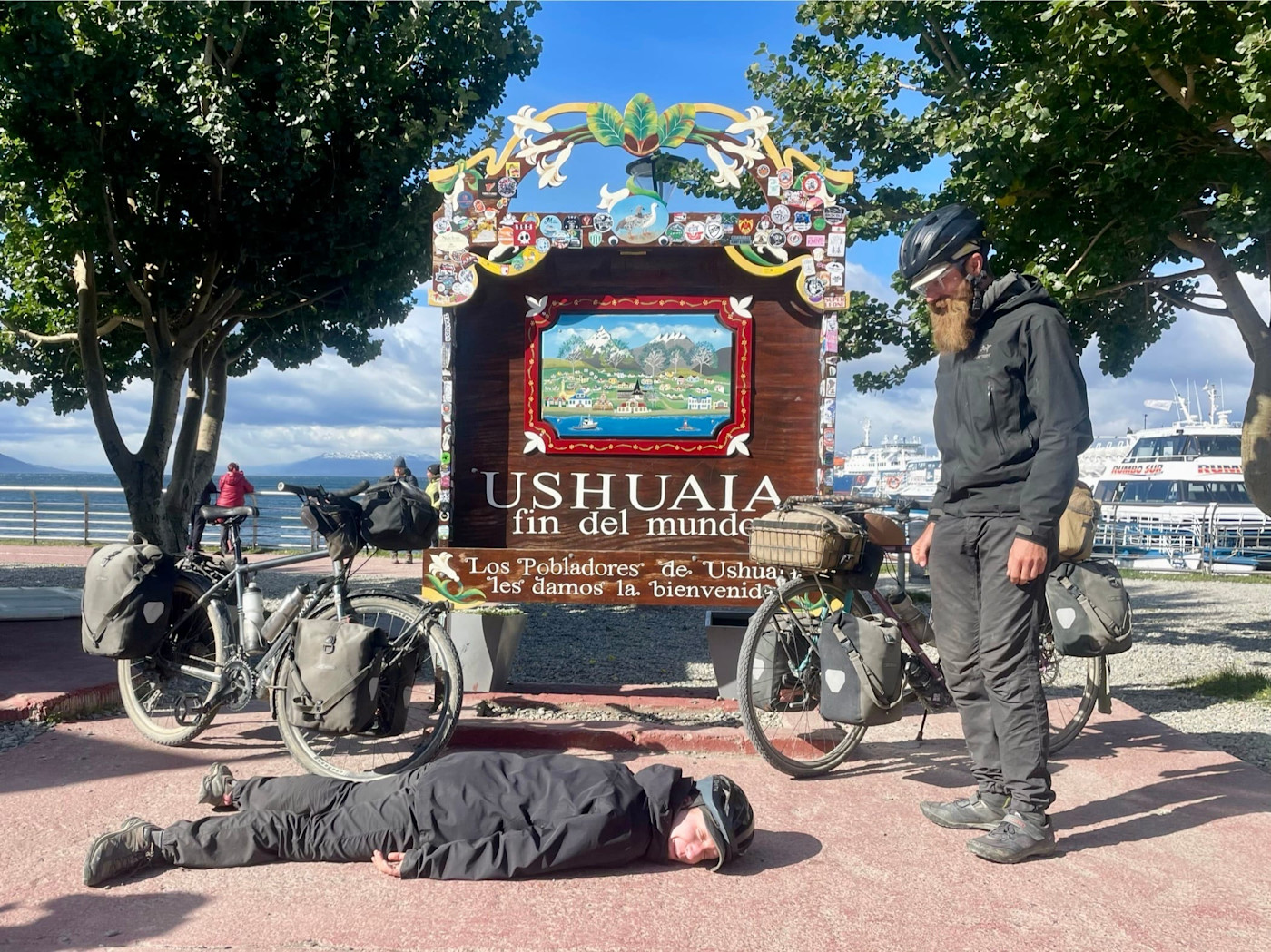 Suzie McCracken이 ‘Ushuaia fin del mundo’라고 적힌 표지판 앞에 누워 있고 Ed Foote가 근처에 서 있다. 짐을 실은 여행용 자전거가 옆에 놓여 있다.