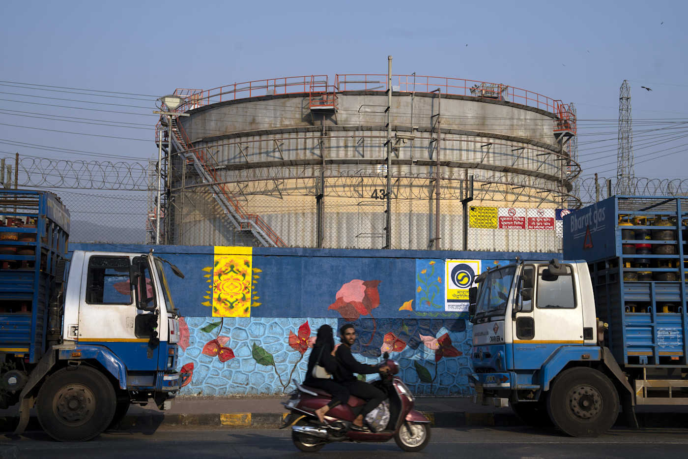 Bharat Petroleum 정유공장의 대형 석유 저장 탱크가 벽화 너머로 보이고, 앞쪽으로 트럭 두 대와 오토바이 한 대가 지나가고 있다.