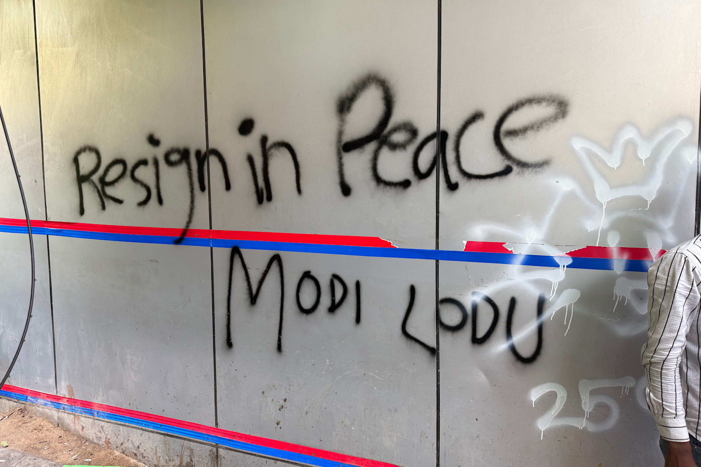 벽에 검은 스프레이 페인트로 “Resign in Peace”와 “Modi Lodu”라고 적힌 그래피티가 있고, 파란색과 빨간색 테이프 선이 붙어 있다.