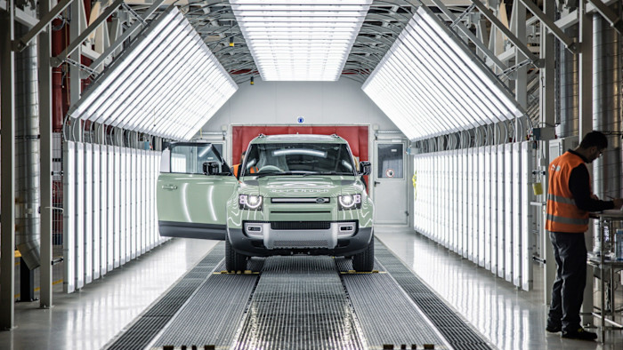 Jaguar Land Rover 공장의 밝은 품질 관리 터널에 Land Rover Defender SUV가 주차돼 있다. 오른쪽에는 안전조끼를 입은 작업자가 클립보드에 무언가를 적고 있다.