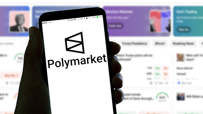손에 든 스마트폰 화면에 Polymarket 로고가 표시되고, 배경에는 Polymarket 앱 인터페이스가 흐릿하게 보인다.