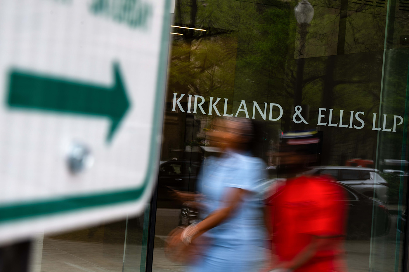 건물 유리 외벽에 표시된 Kirkland & Ellis LLP 간판과 앞을 지나가는 흐릿한 두 사람, 전경의 방향 표지판.