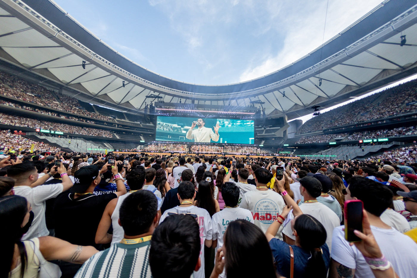 마드리드의 관중으로 가득 찬 경기장에서 배드 버니가 공연하고 있으며, 대형 스크린에 그의 모습이 비치는 가운데 많은 관객이 지켜보며 촬영하고 있다