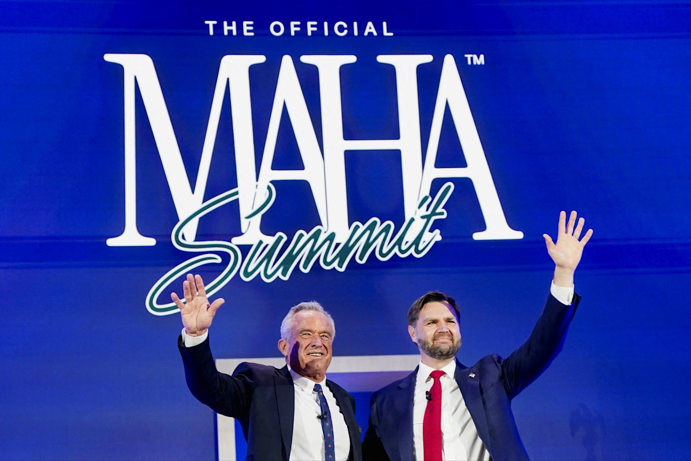 JD 밴스와 로버트 F. 케네디 주니어가 대형 “MAHA 서밋” 표지판 앞 무대에서 손을 흔들고 있다.