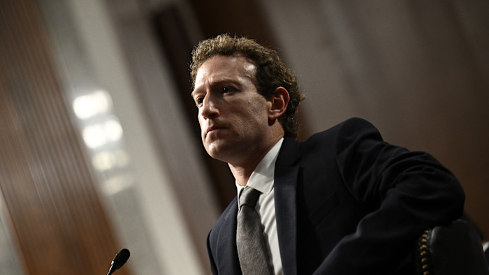 Mark Zuckerberg가 정장을 입고 진지한 표정으로 청문회 테이블에 앉아 있다. 앞에는 마이크가 보인다.