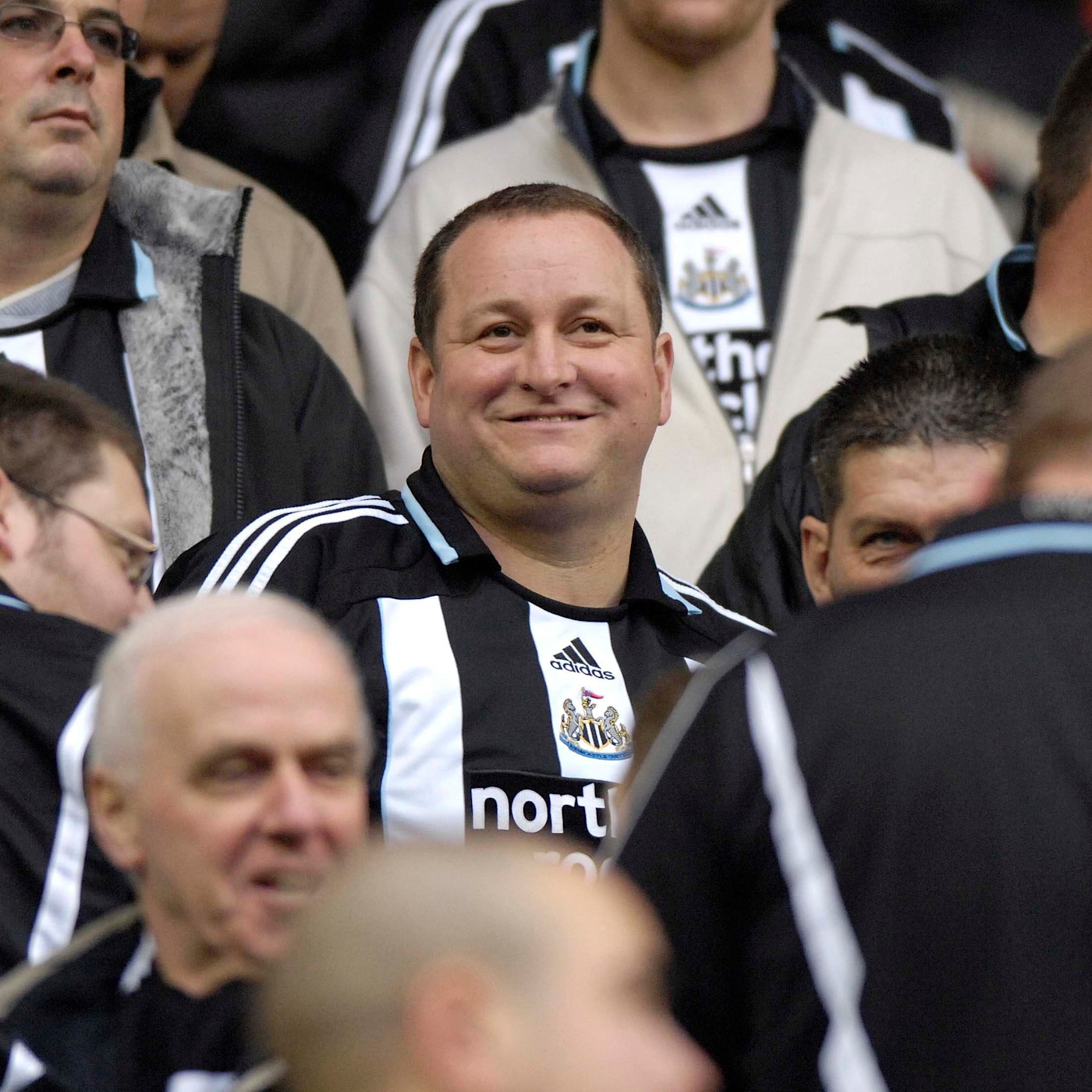 Mike Ashley가 원정석의 Newcastle United 팬들 사이에 서서 미소 지으며 흑백 팀 유니폼을 입고 있다.