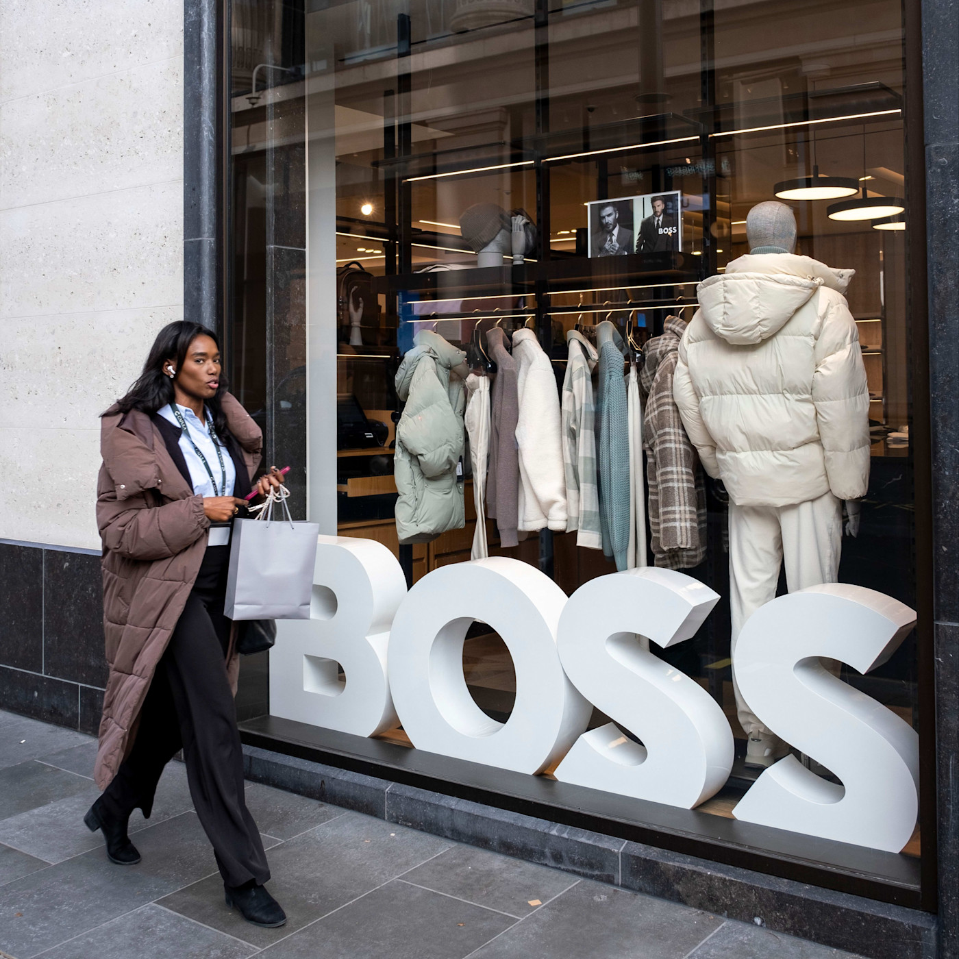 쇼핑백을 든 여성이 겨울 코트와 커다란 BOSS 로고가 전시된 Boss 매장 쇼윈도 앞을 지나가고 있다.