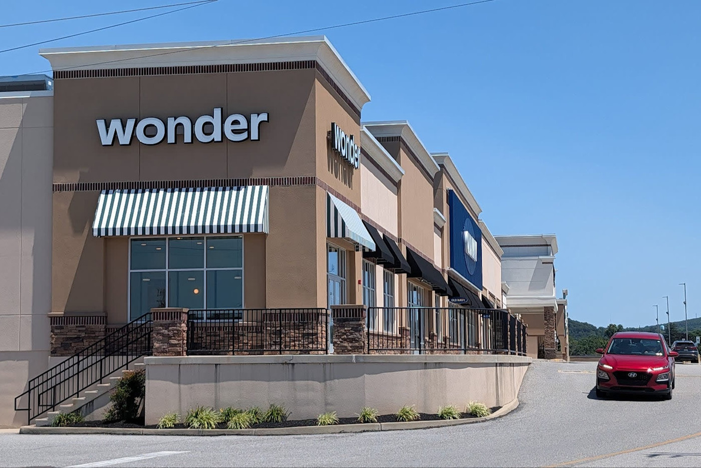 쇼핑센터에 있는 Wonder 매장 전경. 줄무늬 차양 아래로 주차장을 빨간 자동차가 지나가고 있다.