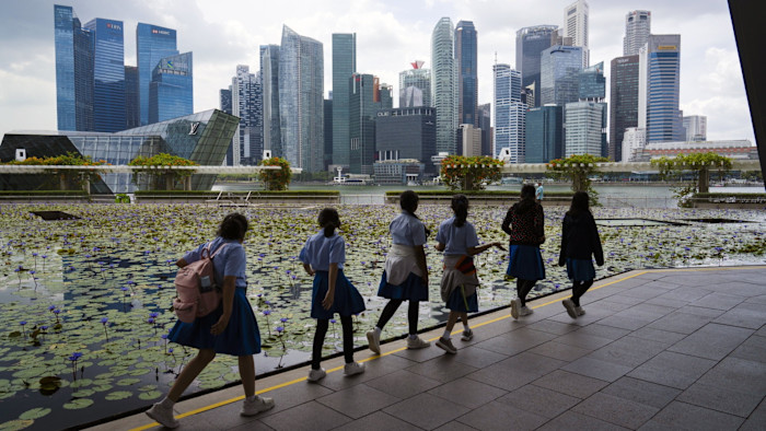 교복을 입은 학생들이 연잎이 떠 있는 연못 옆을 지나가고, 뒤로 Singapore의 스카이라인과 오피스 빌딩이 보인다.