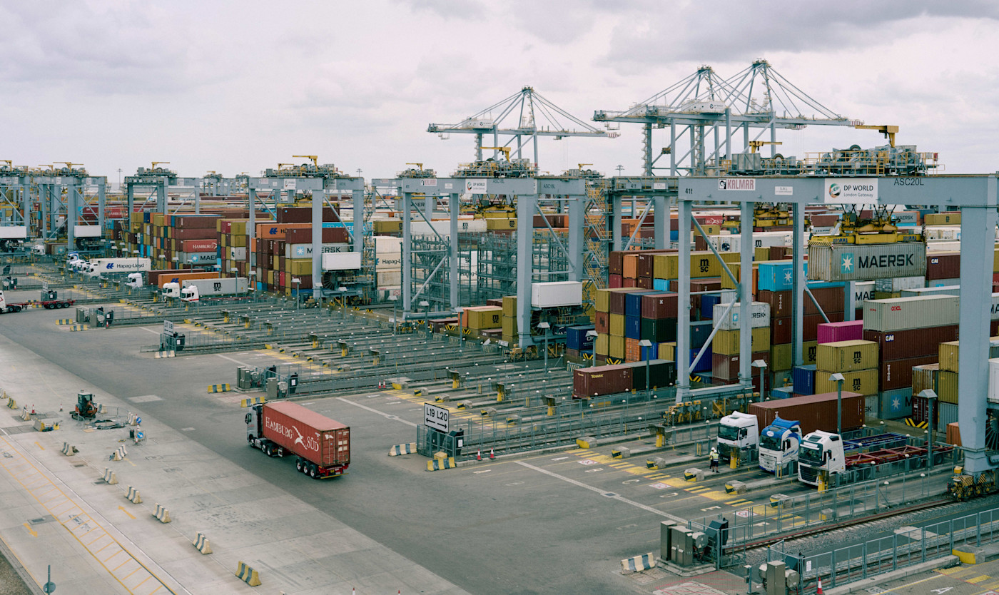 London Gateway 항에서 대형 크레인이 위로 보이는 가운데 줄지어 쌓인 선적 컨테이너. 전경에는 트럭이 보인다.