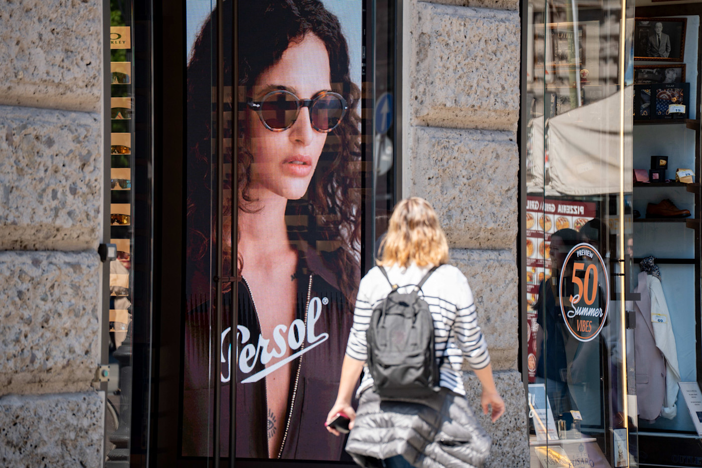 배낭을 멘 여성이 선글라스를 쓴 모델이 등장하는 Persol 아이웨어 대형 광고가 걸린 밀라노의 매장 앞을 지나가고 있다.