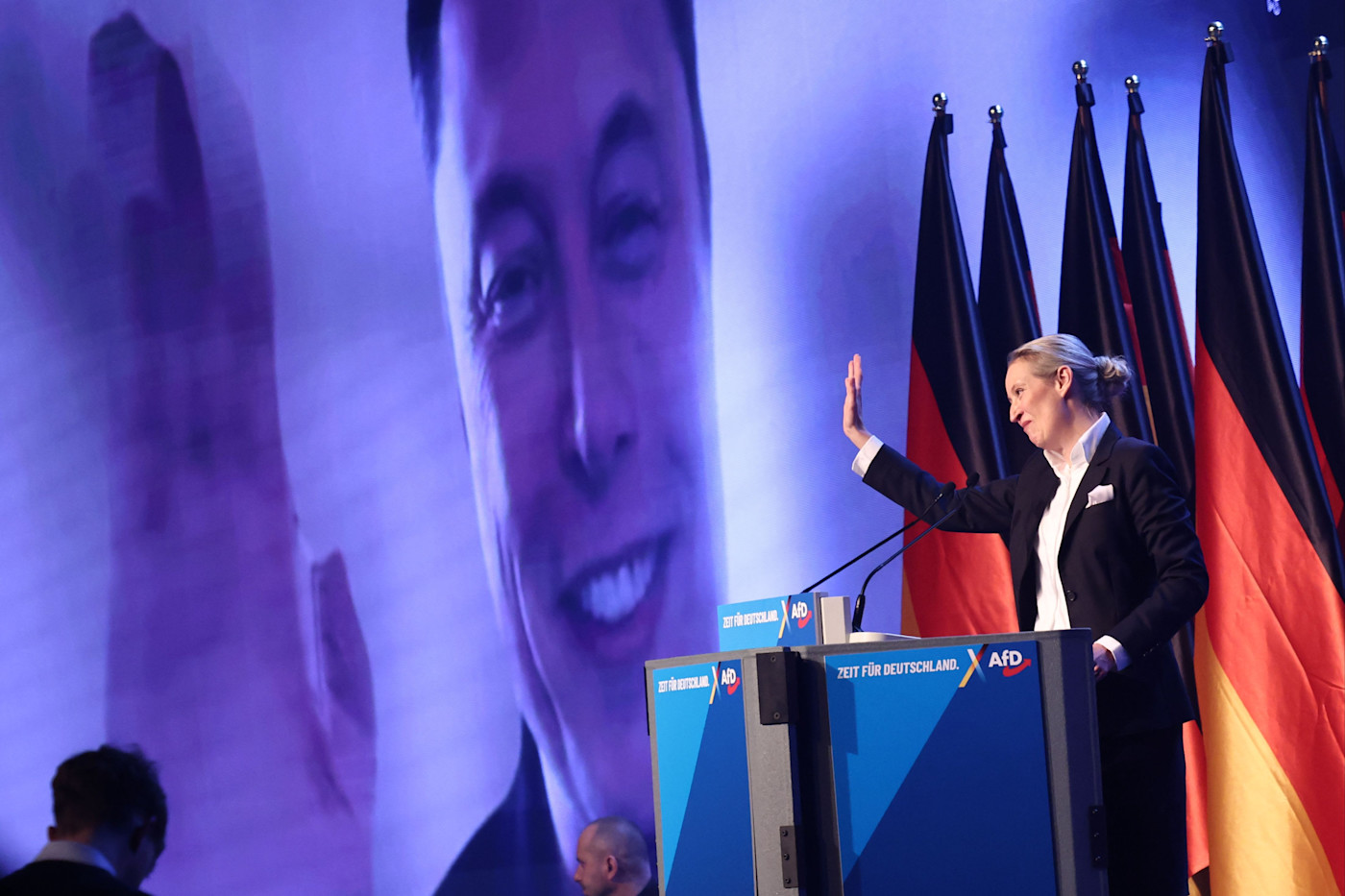 AfD 선거운동 출범 행사 연설에서 Alice Weidel이 뒤편의 독일 국기와 함께 Elon Musk가 나오는 대형 화면을 향해 손짓하고 있다.