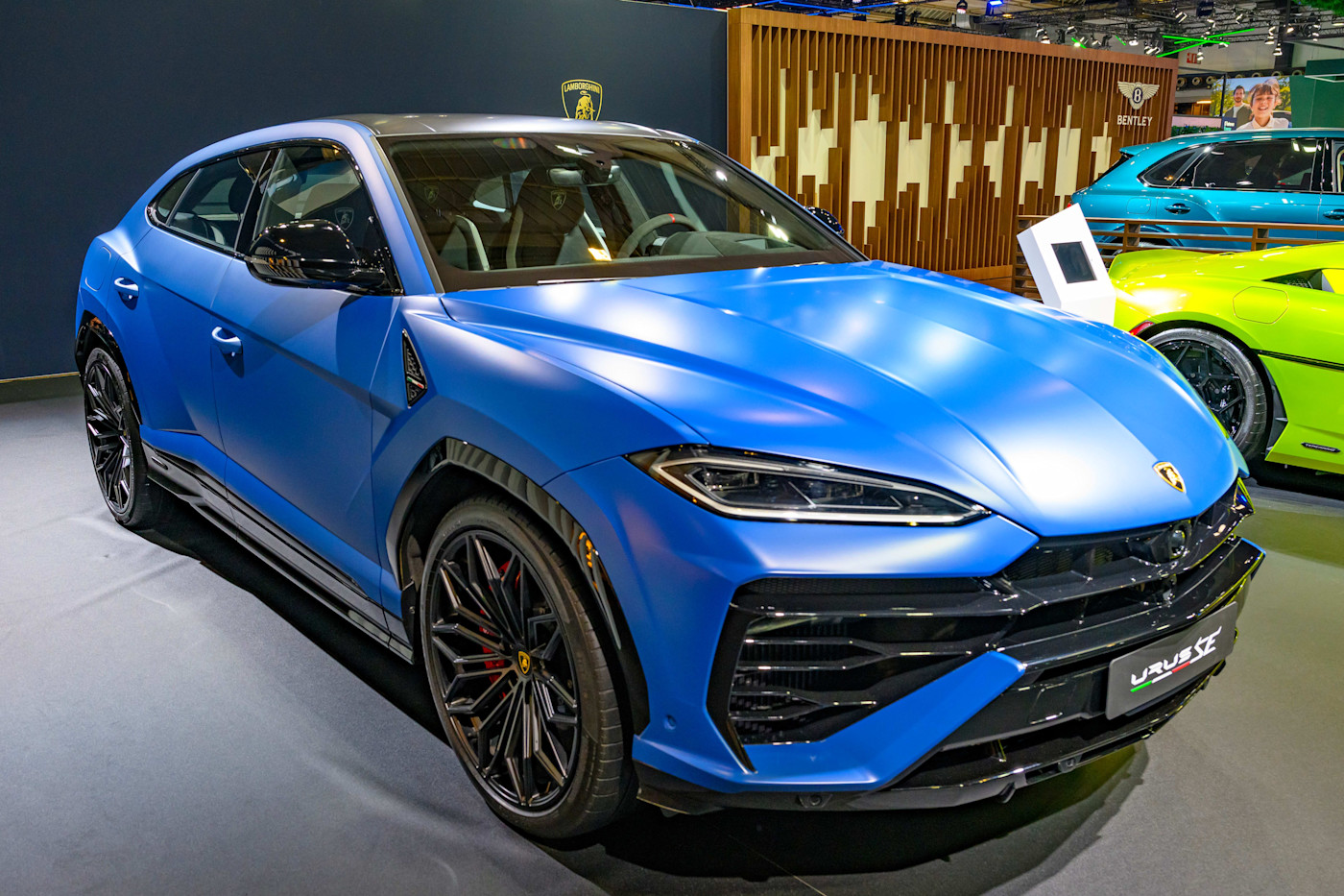 오토쇼에 전시된 무광 블루 Lamborghini Urus SE 퍼포먼스 SUV와 배경에 보이는 다른 럭셔리 차량들.