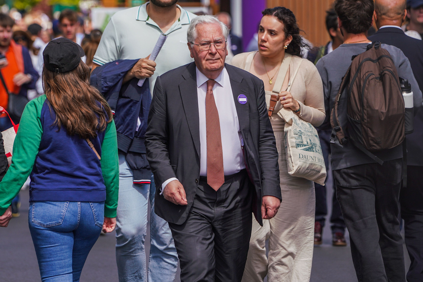 Mervyn King이 정장과 주황색 넥타이를 착용하고 재킷에 보라색 행사 배지를 단 채 Wimbledon의 군중 사이를 걷고 있다.