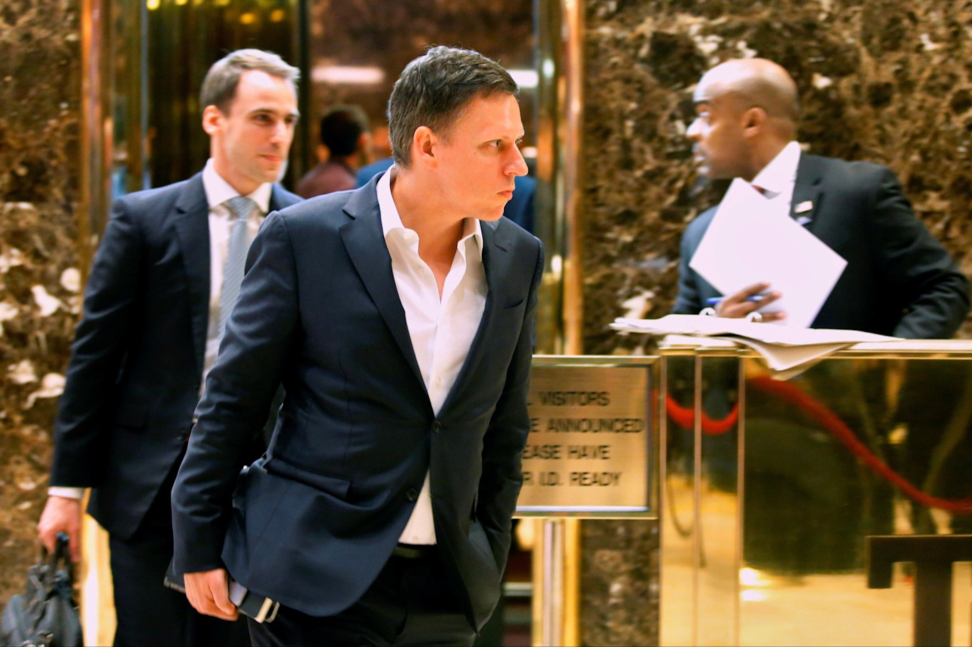 정장을 입고 휴대전화를 든 Peter Thiel이 Trump Tower 엘리베이터에서 내리고 있으며, 근처에 정장 차림의 남성 두 명이 있다.