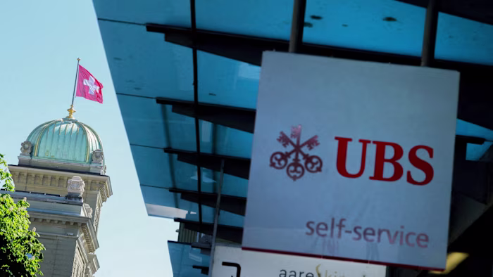전경에 UBS 로고 간판이 있고 배경에 스위스 의회 건물과 스위스 국기가 보이는 모습.