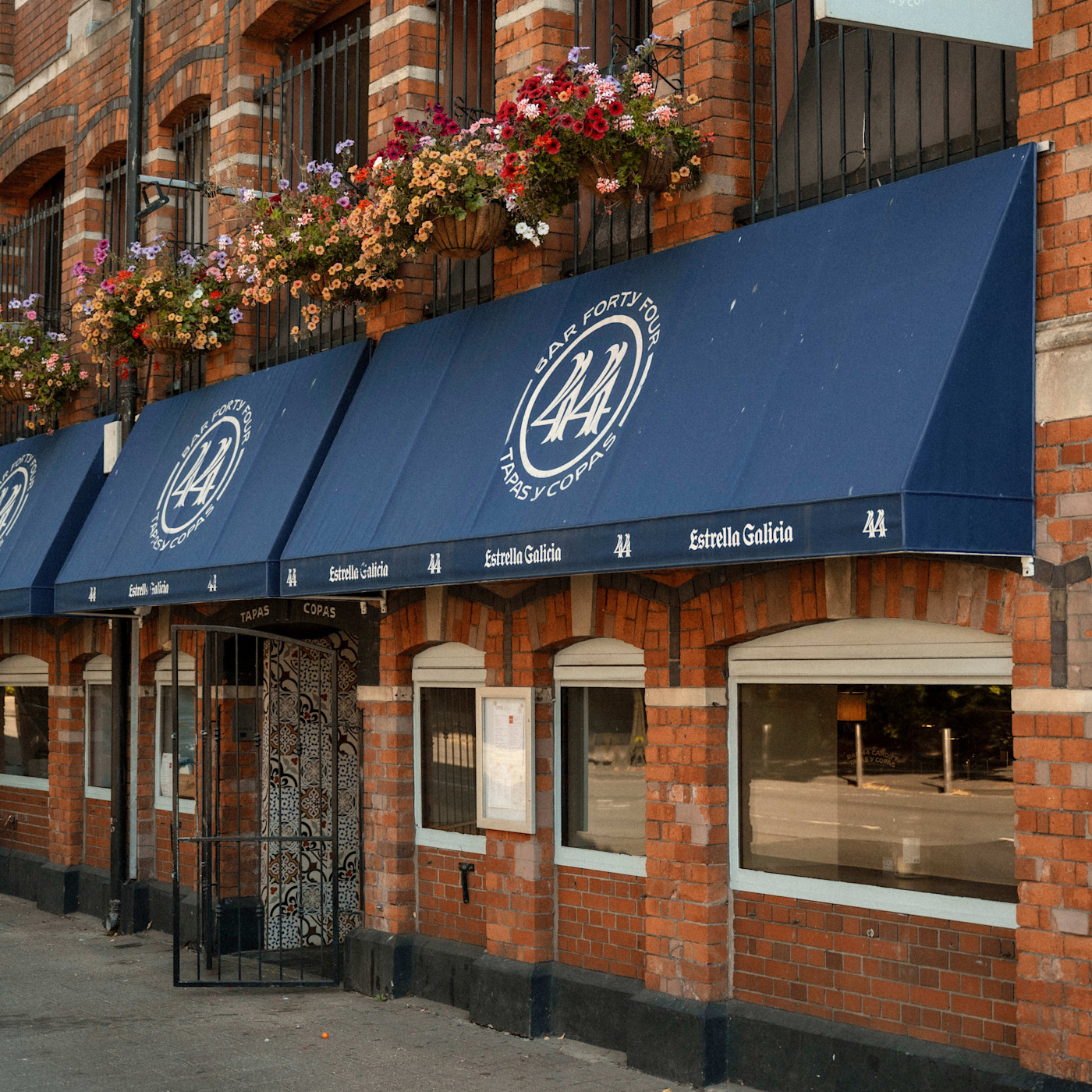 Westgate Street의 Bar 44 입구 위에 ‘Bar 44 Tapas y Copas’ 로고가 있는 파란 차양과 매달린 꽃.