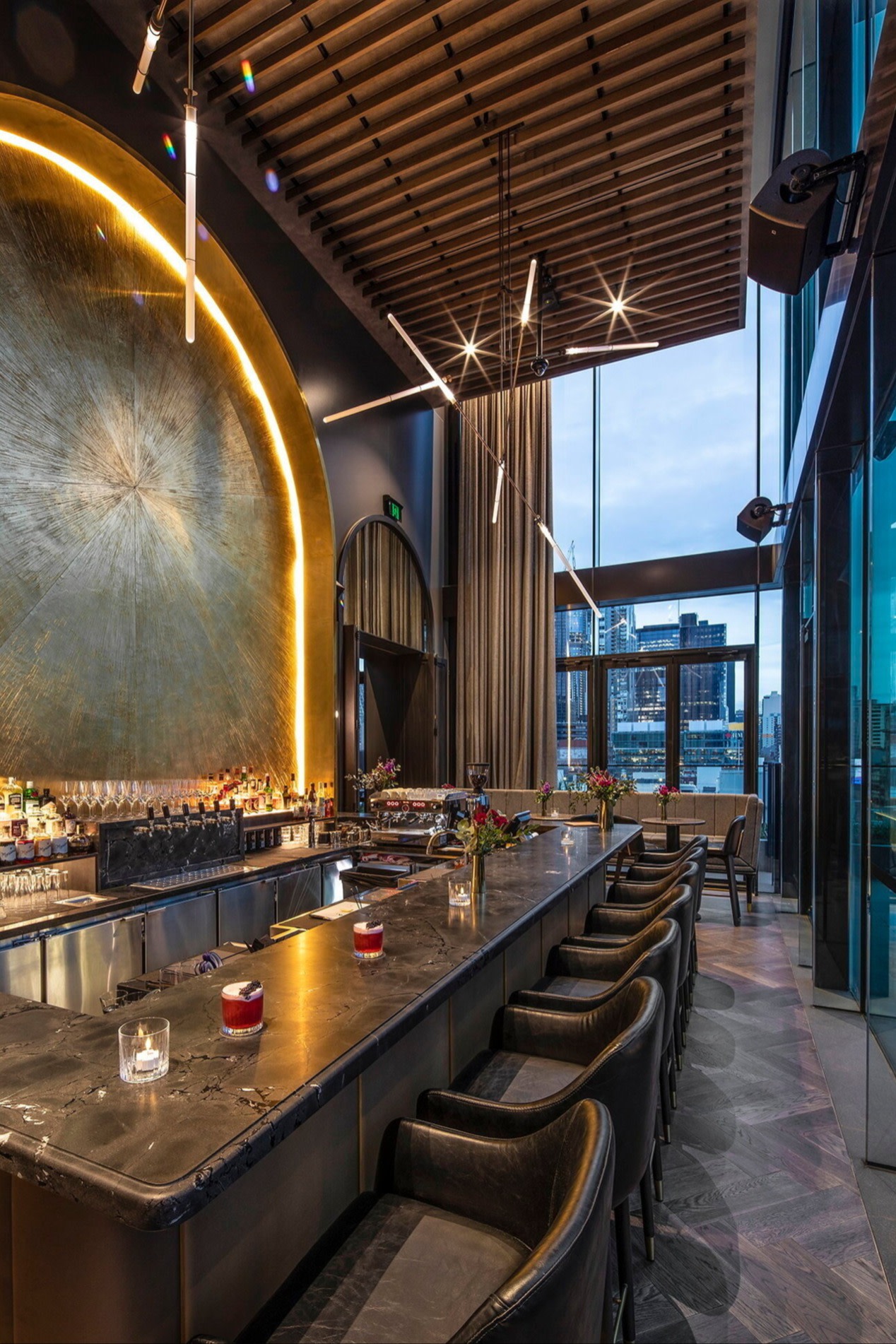 Blacksmith Bar & Grill 내부 전경. 긴 대리석 카운터와 검은색 가죽 스툴, 바 뒤편의 크고 질감 있는 금색 벽 장식이 보인다. 바닥부터 천장까지 이어지는 창밖으로 도시 풍경이 펼쳐진다.