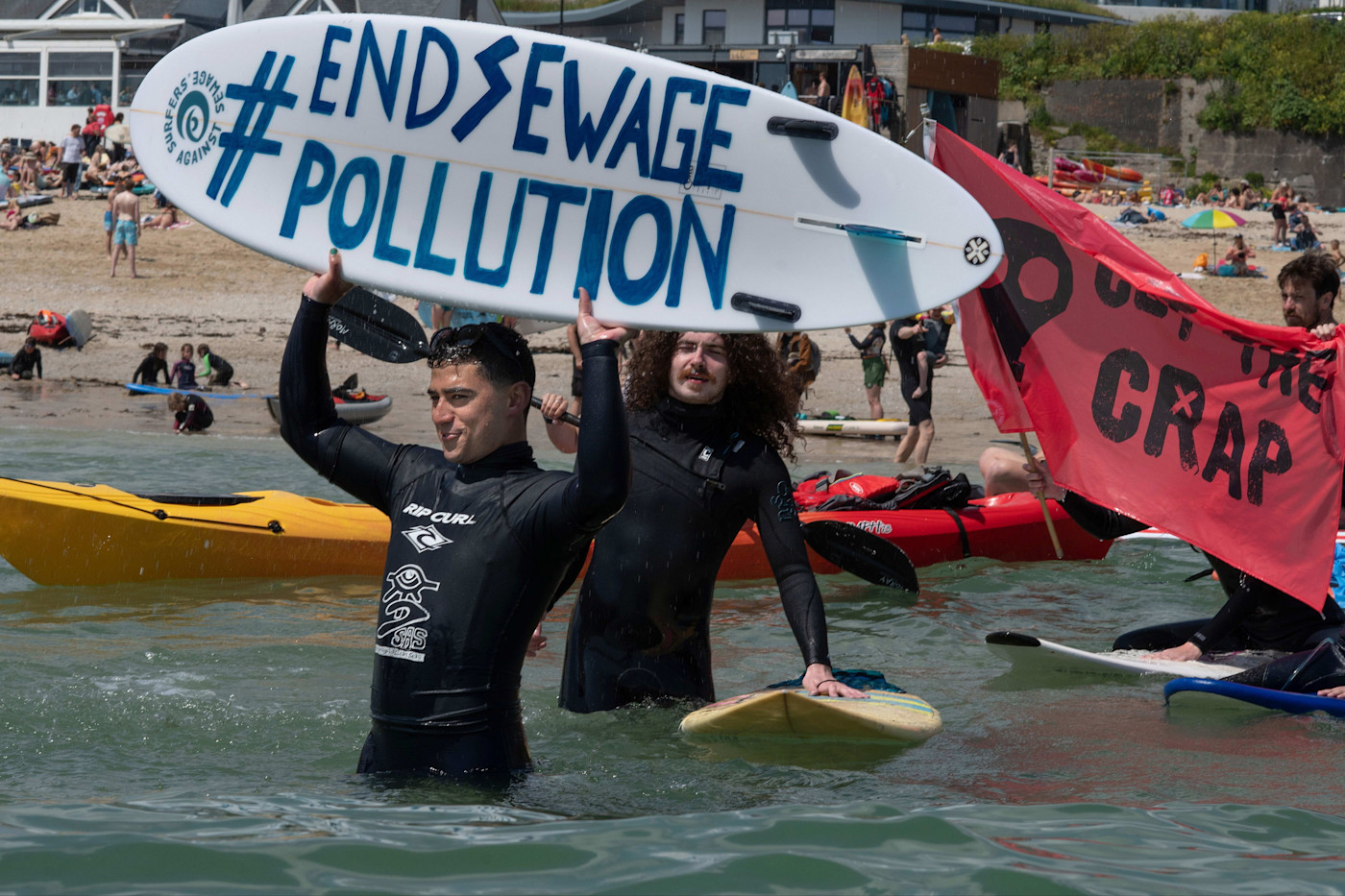 시위 중 바다에 선 잠수복 차림의 사람들이 “#EndSewagePollution”이라고 적힌 서프보드와 “Cut the Crap”이라고 적힌 빨간 깃발을 들고 있다.