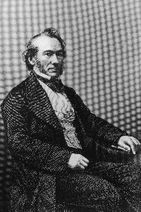 격식 있는 19세기 복장을 하고 정면을 바라보며 한 손을 무릎에 올린 채 앉아 있는 Richard Cobden.