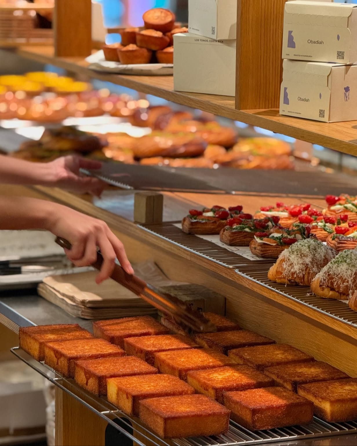 Arôme Bakery에서 한 사람이 집게로 골든 허니 토스트 한 조각을 집어 드는 동안, 식힘망 위에 황금빛 허니 토스트 조각들이 줄지어 놓여 있고 뒤쪽에는 다양한 페이스트리가 보인다.