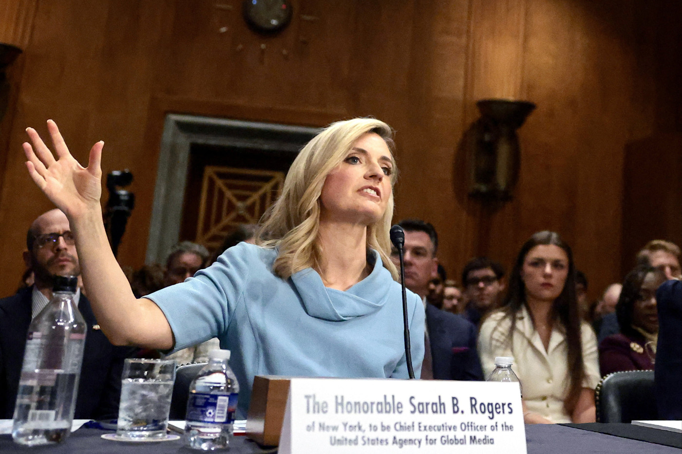 Sarah Rogers가 상원 위원회 청문회에서 이름표 뒤에 앉아 증언하며 손짓하고 있고, 배경에는 방청객이 보인다.