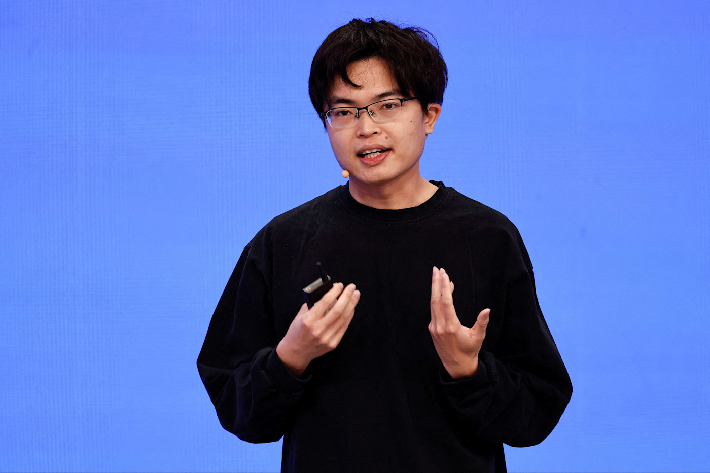 양즈린이 기기를 들고 단색 파란색 배경 앞에 서서 연설하고 있다.