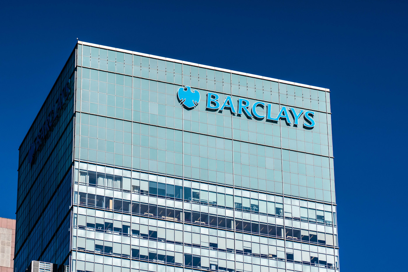 맑은 푸른 하늘 아래 유리 오피스 빌딩 상층부에 표시된 Barclays 로고와 사명