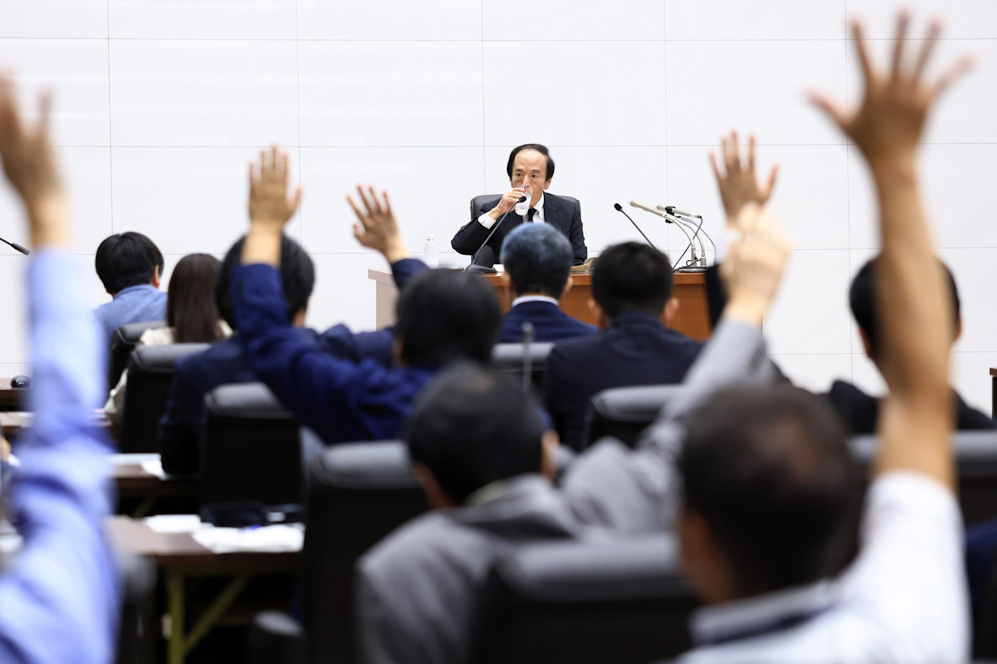 기자회견에서 우에다 카즈오가 연단에 앉아 물을 마시는 동안 객석의 여러 사람이 손을 들고 있다.