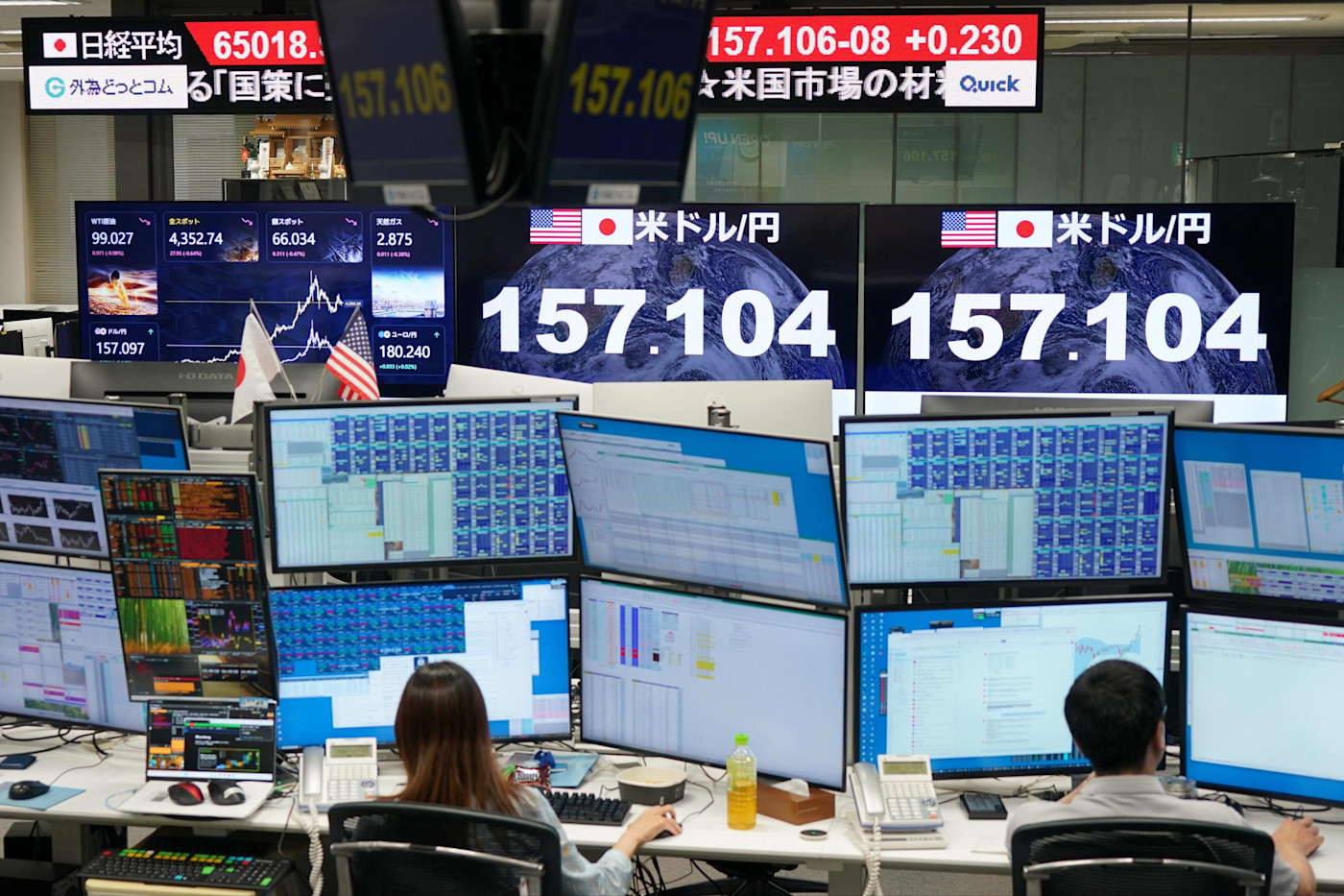 전광판에는 엔/달러 환율 157.104가 표시되어 있고, 거래실에서 사람들이 컴퓨터 단말기 앞에서 일하고 있다.