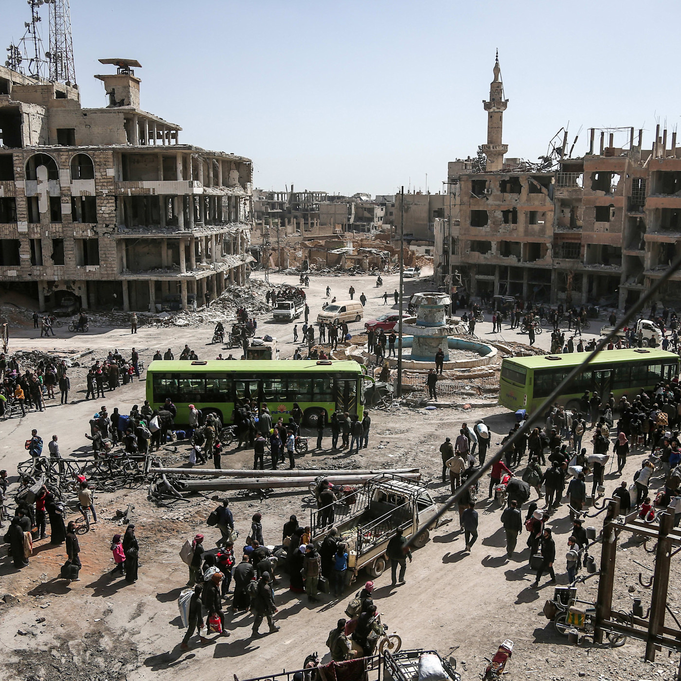 Crowds of civilians and rebel fighters gather near green buses amid damaged buildings in Arbin during an evacuation.