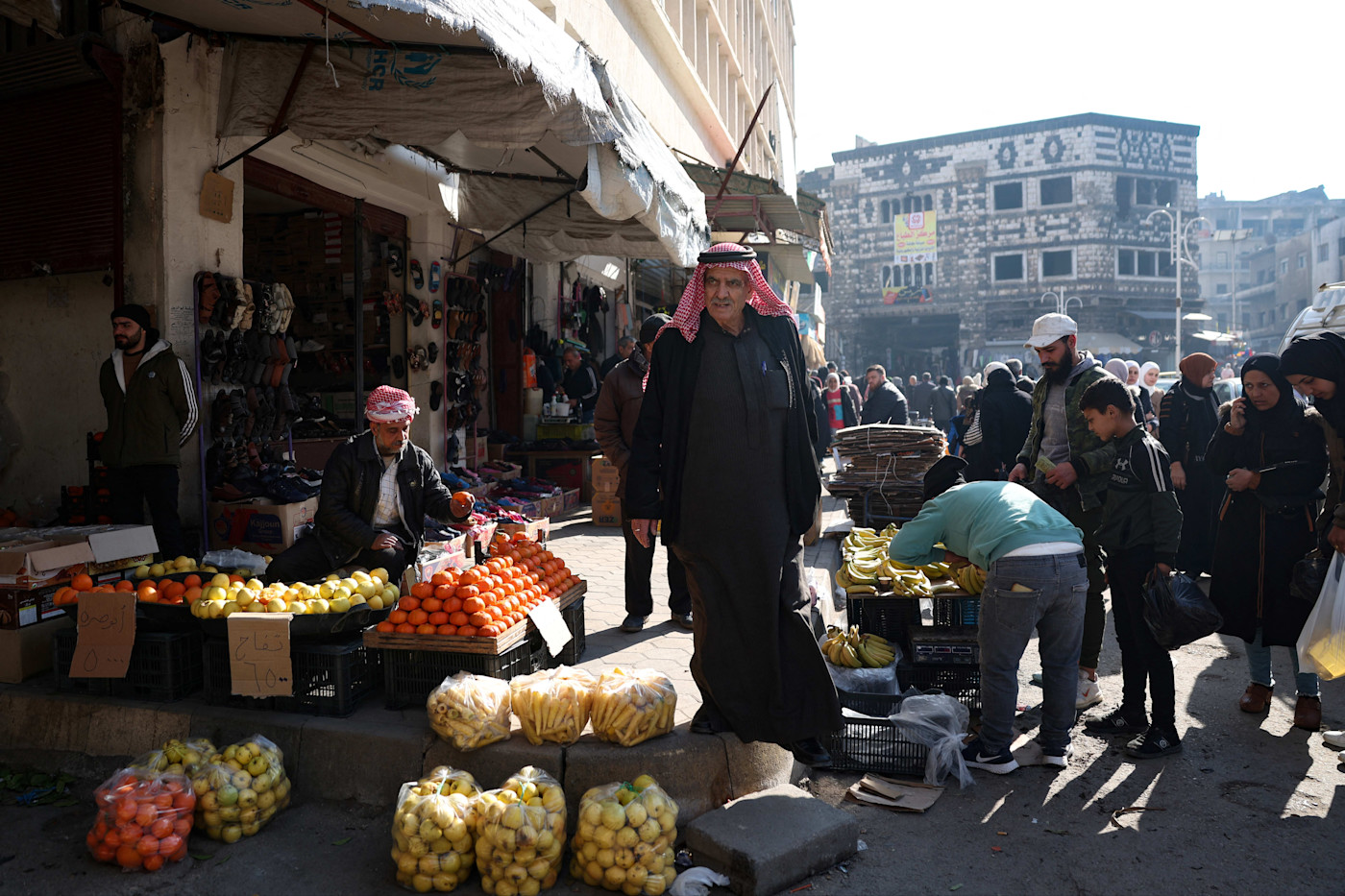 A fruit vendor arranges oranges and apples at a busy outdoor market in Homs, with shoppers browsing nearby stalls.