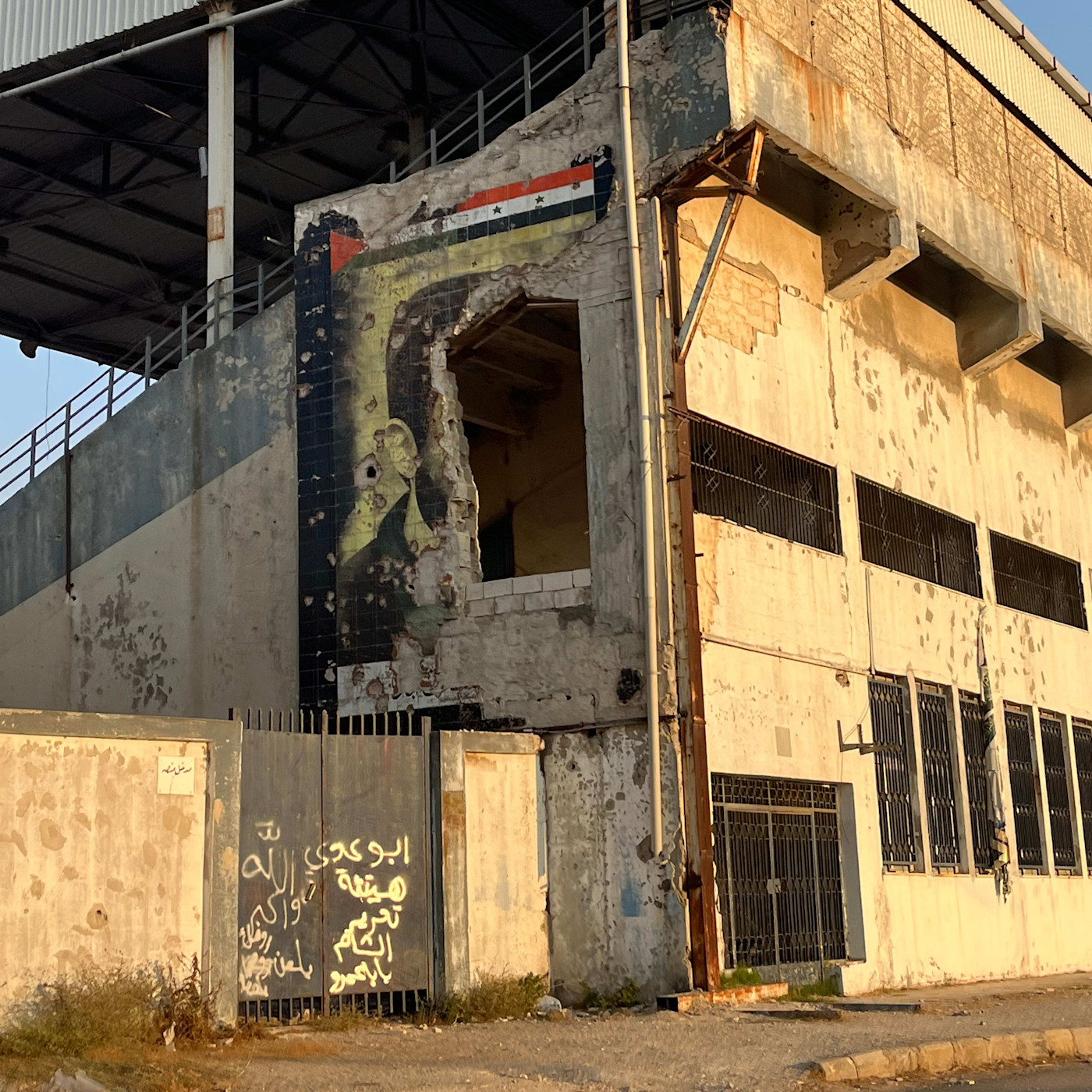 A partially destroyed mural of Bashar al-Assad is visible on a damaged building, with the Syrian flag painted above it.