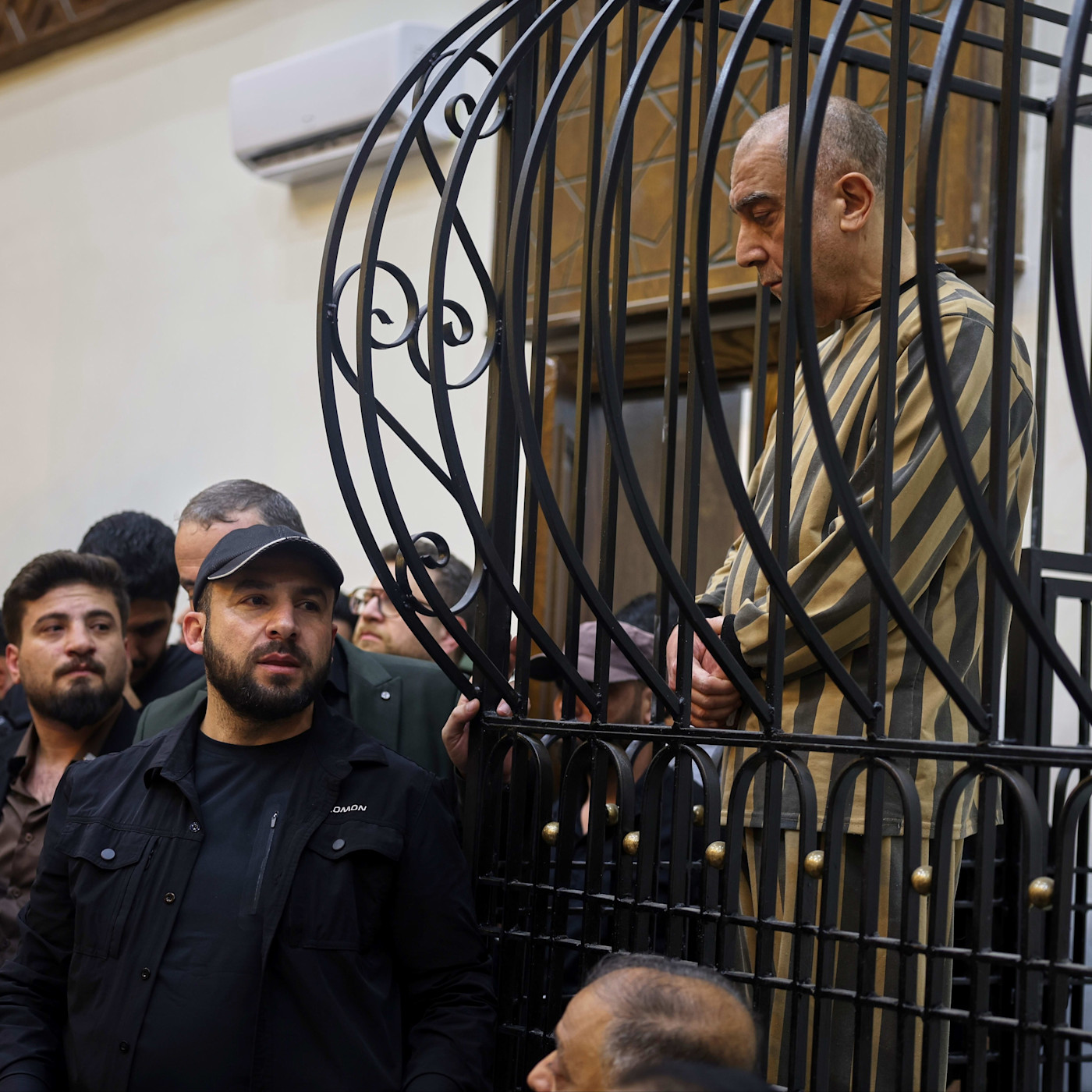 Atef Najib stands inside a metal defendants' cage during a trial session, wearing a striped prison uniform, with several people nearby.